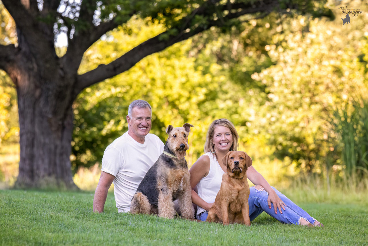 A man, woman, an Airedale and Red Fox Lab sit in the grass at Johnson Park in Walker, MI.